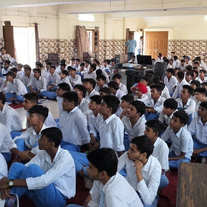 A long school hall filled with students in white uniforms seated on the floor, facing a presentation at the far end.