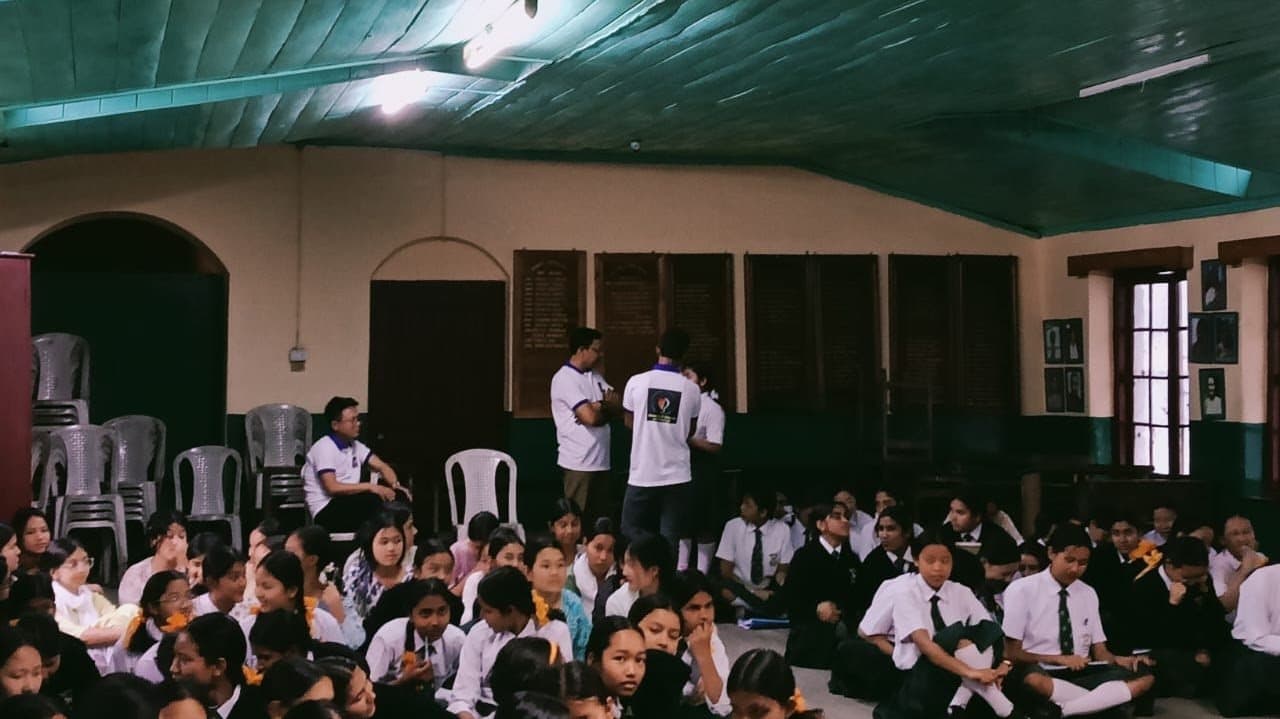 Students seated on the floor of a hall with a green roof, facilitators standing at the back.