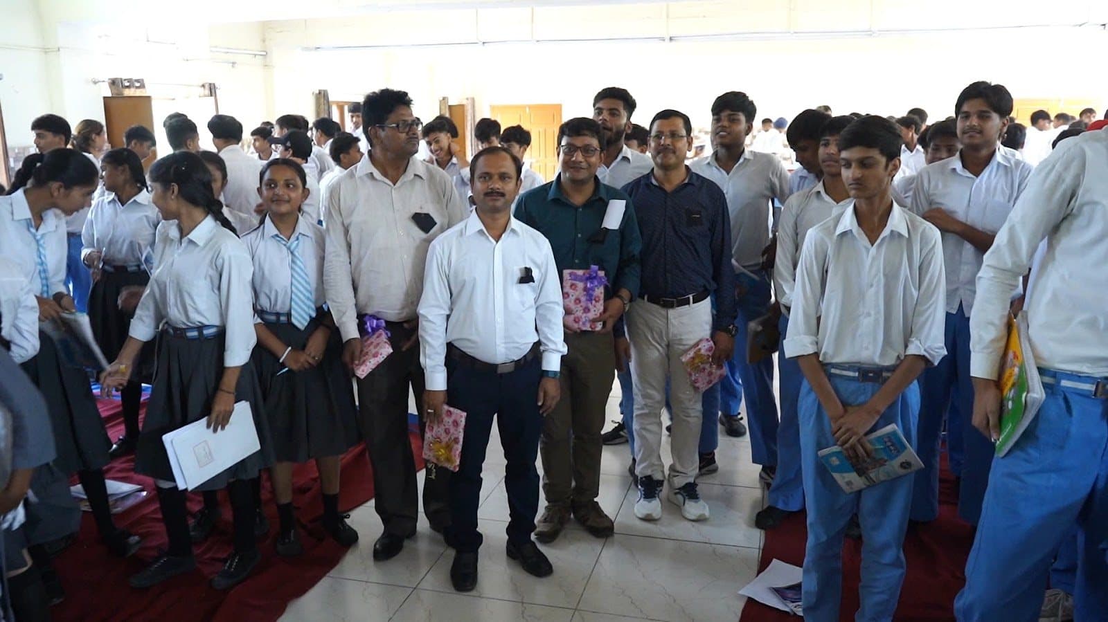 A crowd of students in uniform and adults standing together in a school hall, several holding booklets.