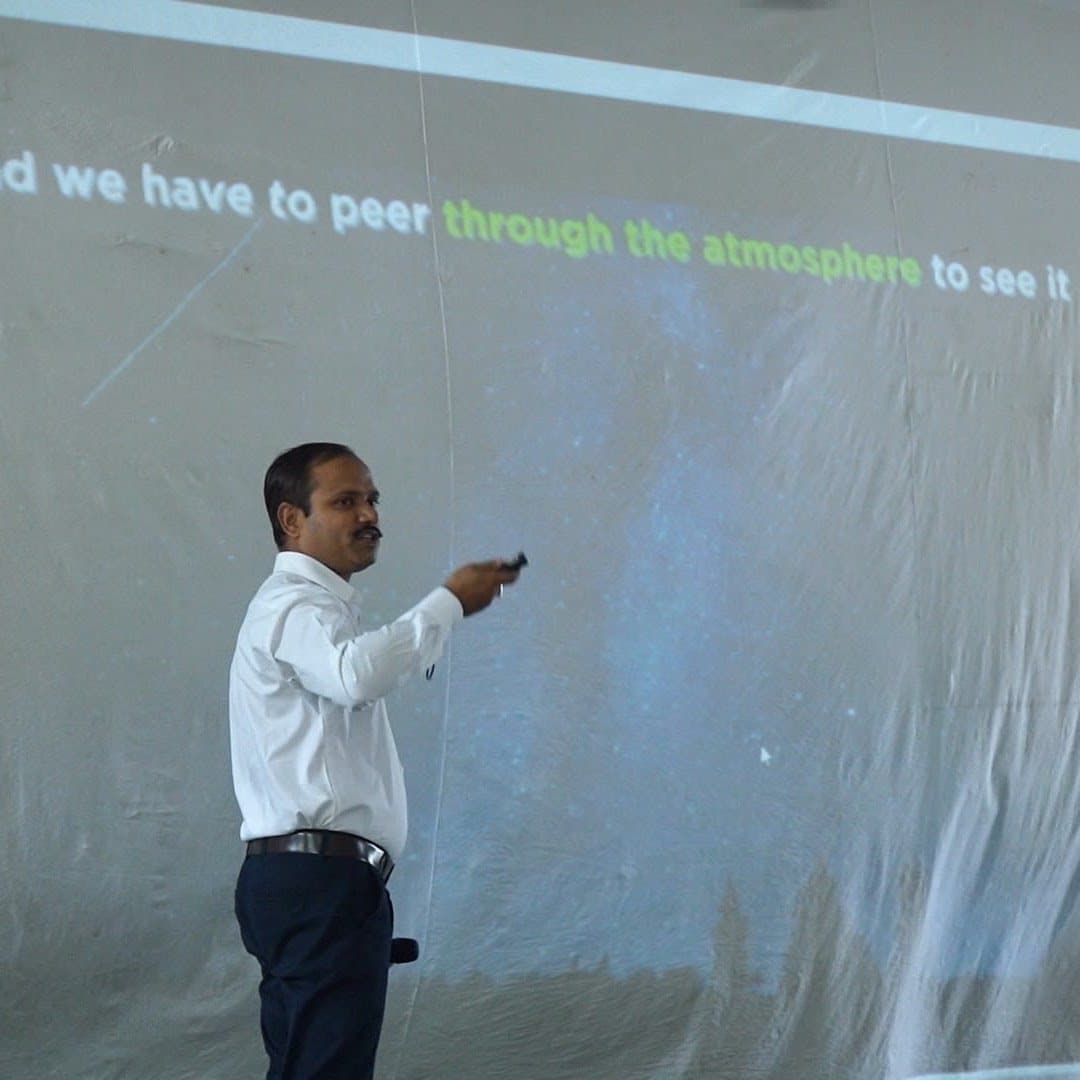 A speaker pointing at a large projected slide reading “and we have to peer through the atmosphere to see it”.