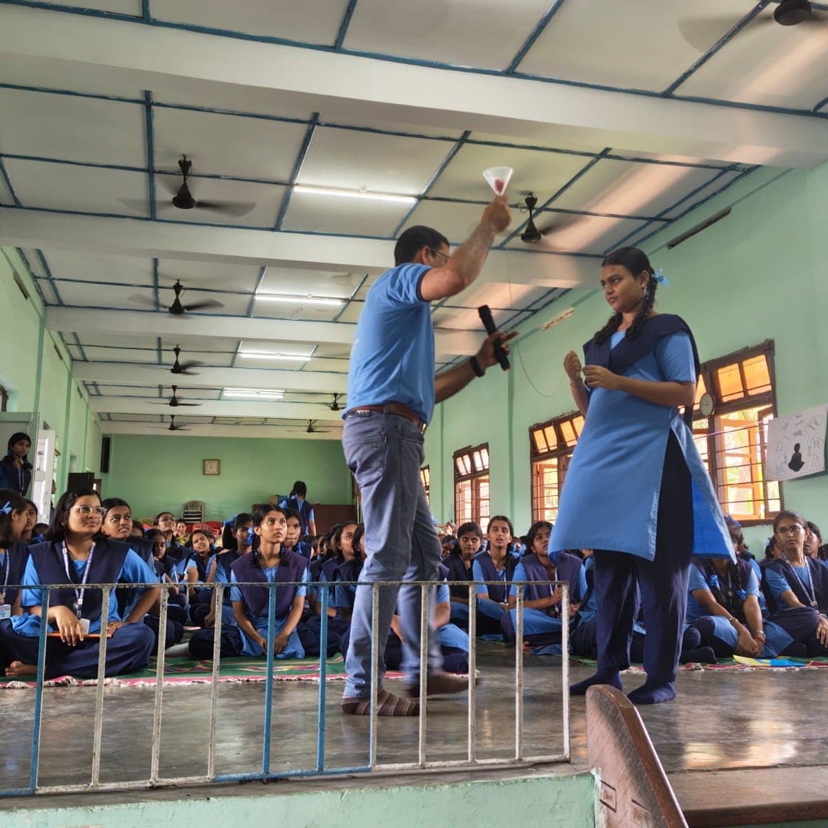 A facilitator holding a funnel aloft beside a teacher, with students watching from behind a railing.