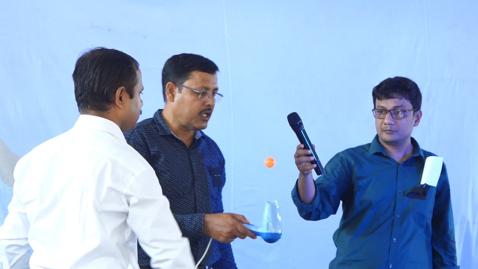 Three people at the front of a room, one holding an orange ball suspended in mid-air above an upturned funnel.