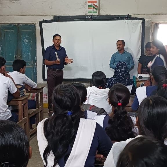 A facilitator addressing students seated at desks in a classroom, a projector screen behind him.