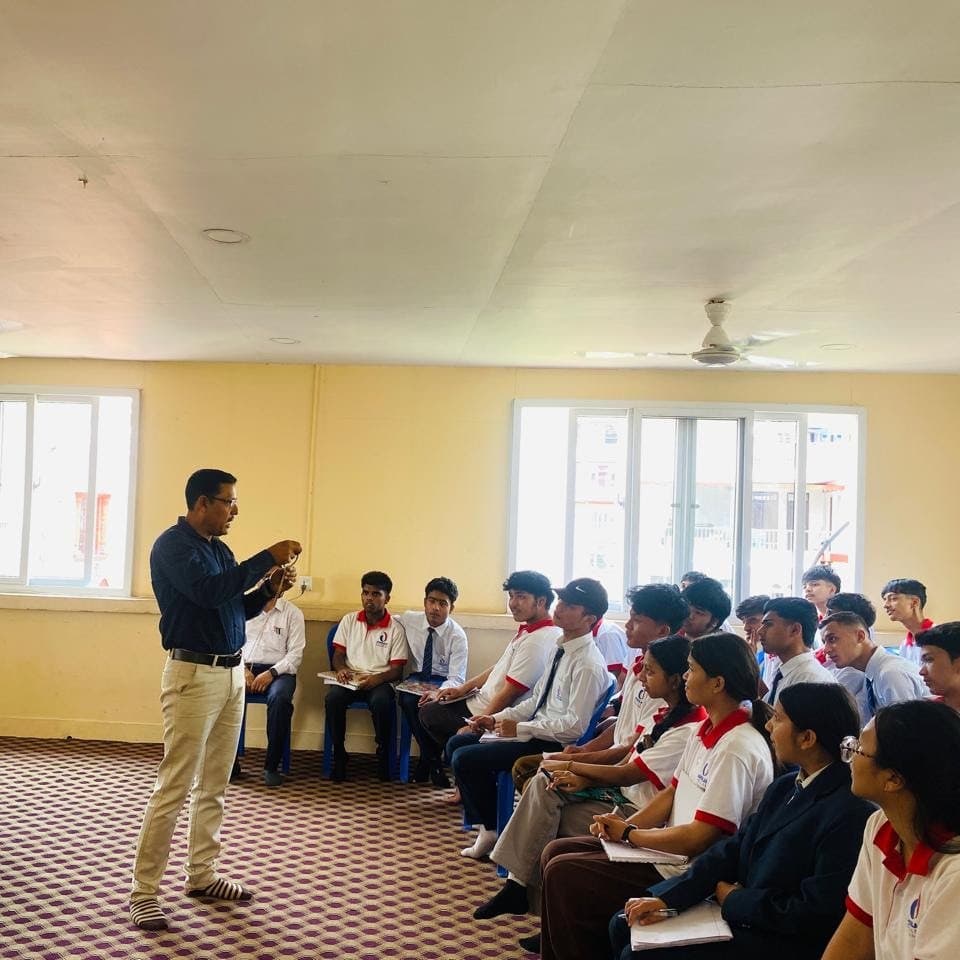 A facilitator addressing students in blazers seated along the walls of a yellow-walled room.