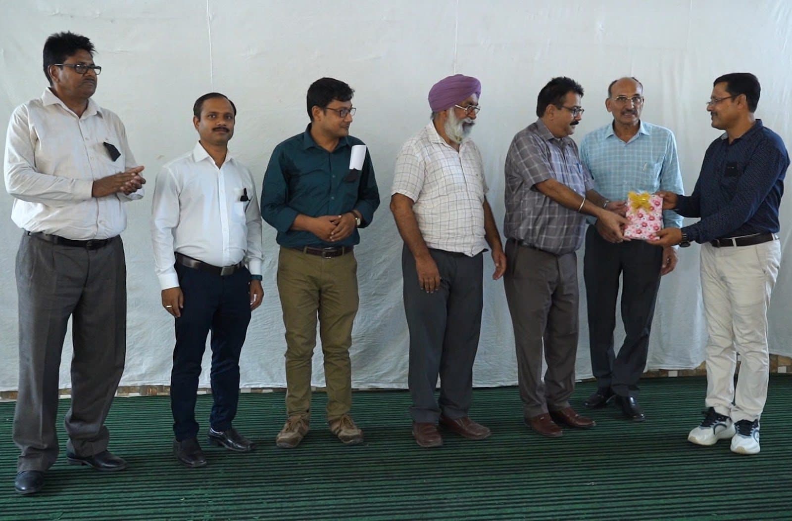 A line of men across a stage during a closing presentation, one holding a wrapped gift.