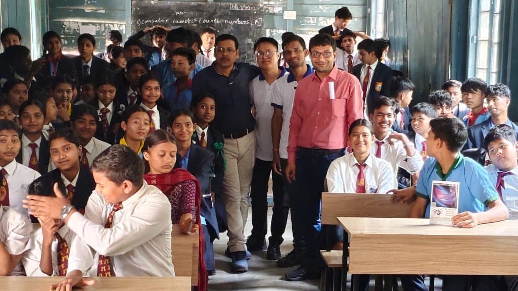 Students crowded together in a classroom, some seated at a desk at the front.