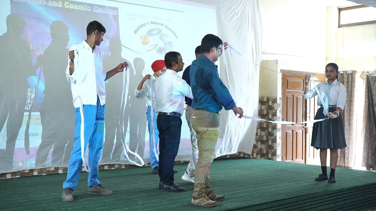 Students and facilitators on a stage holding lengths of white paper, a projected slide behind.