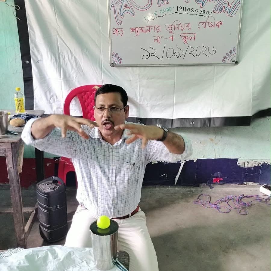 A facilitator gesturing with both hands mid-explanation, with apparatus and a ball on the table in front of him.