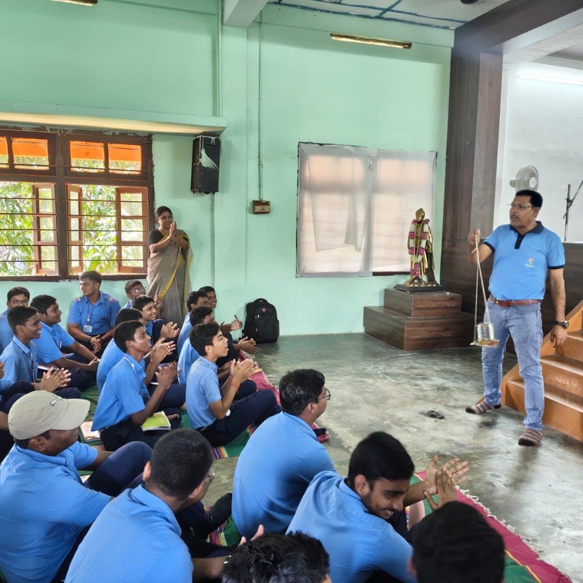 Students in blue uniforms seated on the floor of a hall as a facilitator speaks from the front.