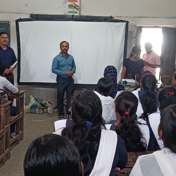 Students seated at desks facing a projector screen, two facilitators standing at the front of the classroom.