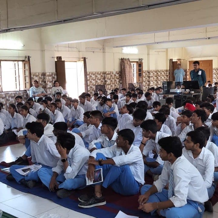 Students in white uniforms seated on the floor of a bright hall, watching the front.
