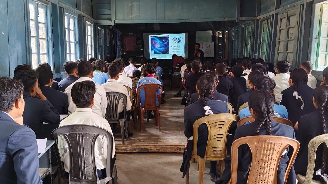 An audience seated on plastic chairs facing a projector screen in a school hall.