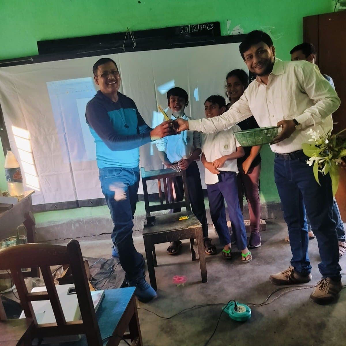 Facilitators handing apparatus to students in a classroom with a green wall and a projector running.