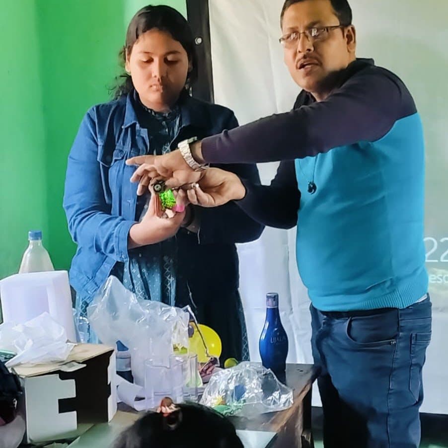 A facilitator guiding a student's hands through a demonstration, a table of bottles and materials beside them.