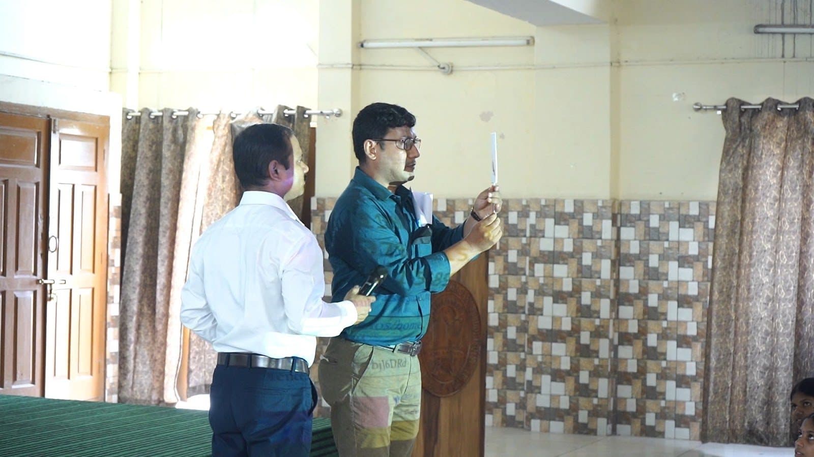 Two facilitators at the front of a hall, one holding a small object up to the light.