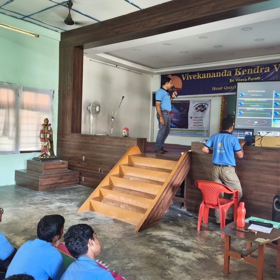 A school hall with a wooden staircase, students in blue uniforms seated as a session runs at the front.