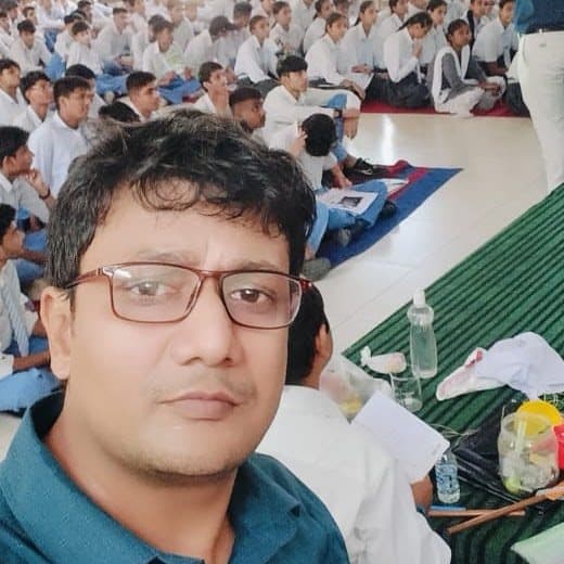 A close-up photograph of a facilitator with a hall of seated students visible behind him.