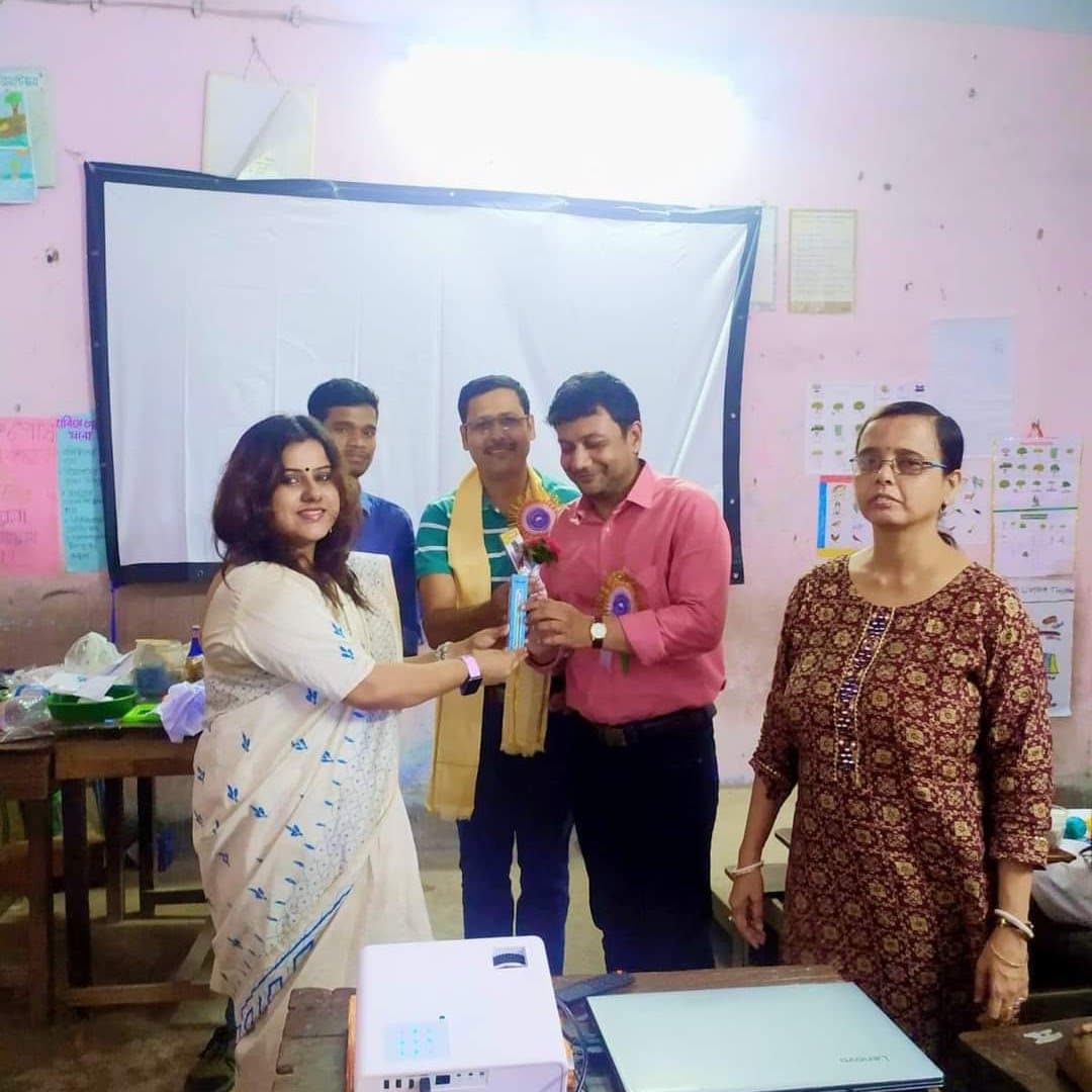 A teacher presenting a memento to a visiting facilitator in a classroom, colleagues standing alongside.