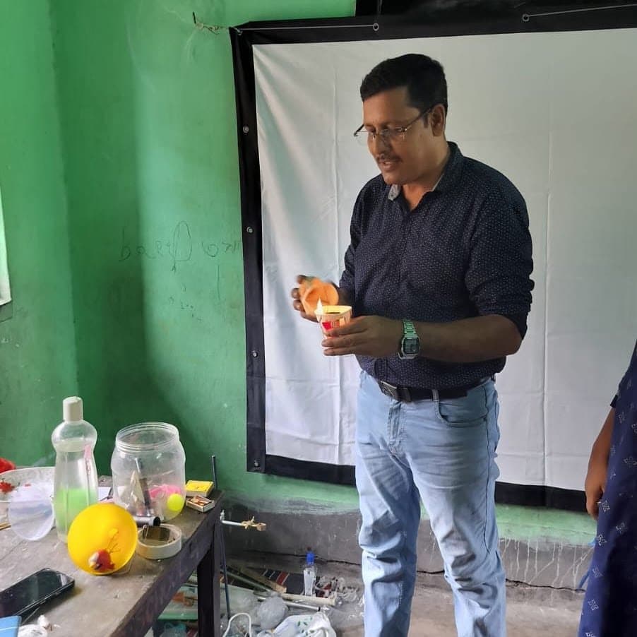 A facilitator holding up an orange ball in front of a screen, a table of apparatus beside him.