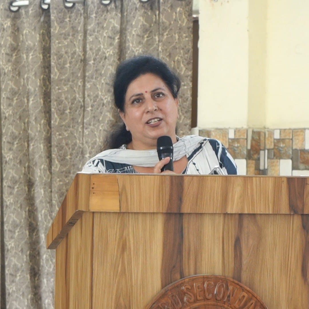 A teacher speaking into a microphone from a wooden lectern at the front of a hall.