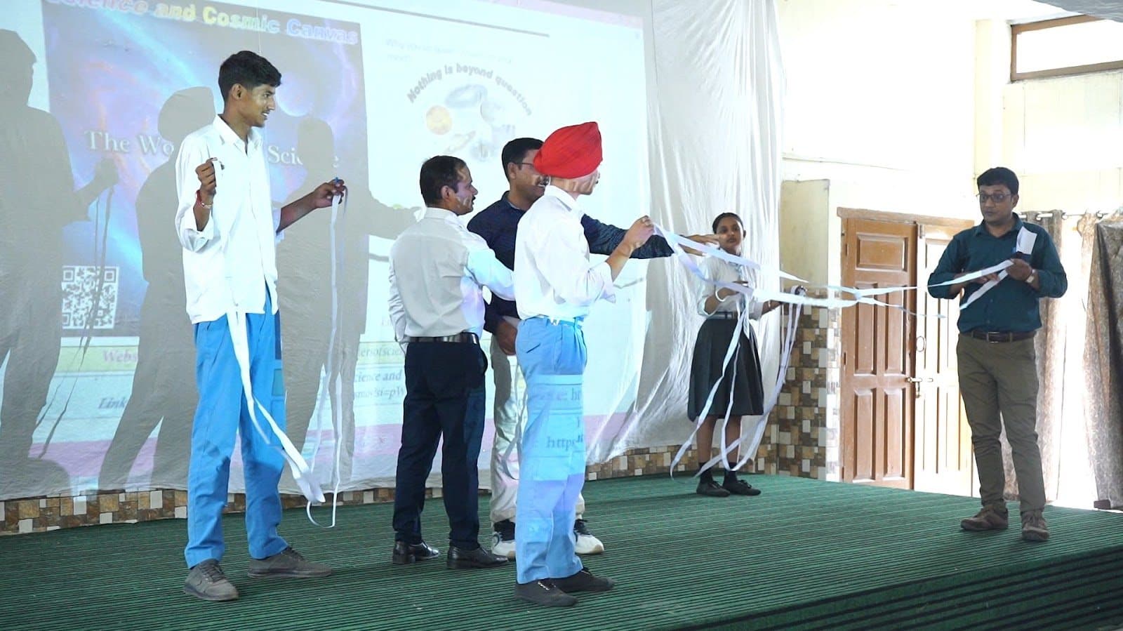 Students on a stage holding long white strips while a facilitator explains, a projected slide behind them.