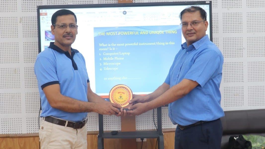 Two men holding a commemorative plaque between them in front of a screen showing a question slide.