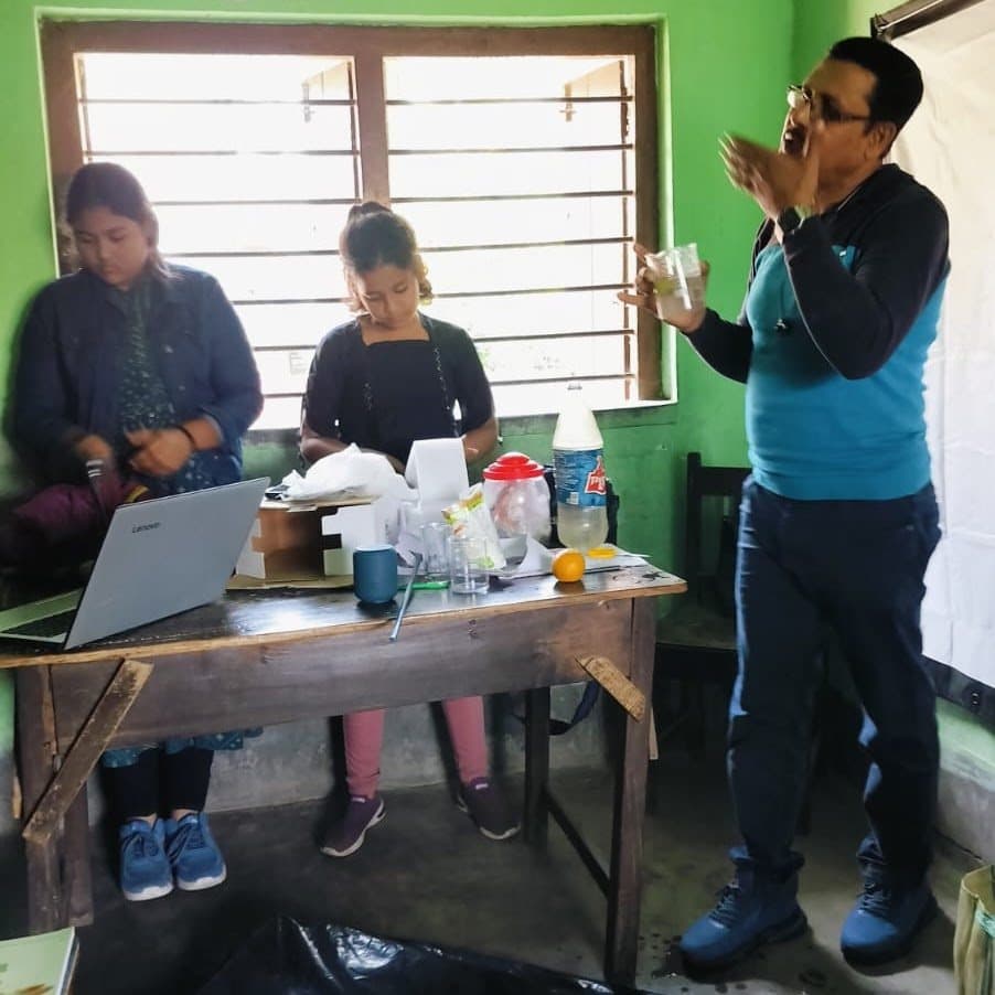 Two young women preparing apparatus on a table while a facilitator explains, beside a window.