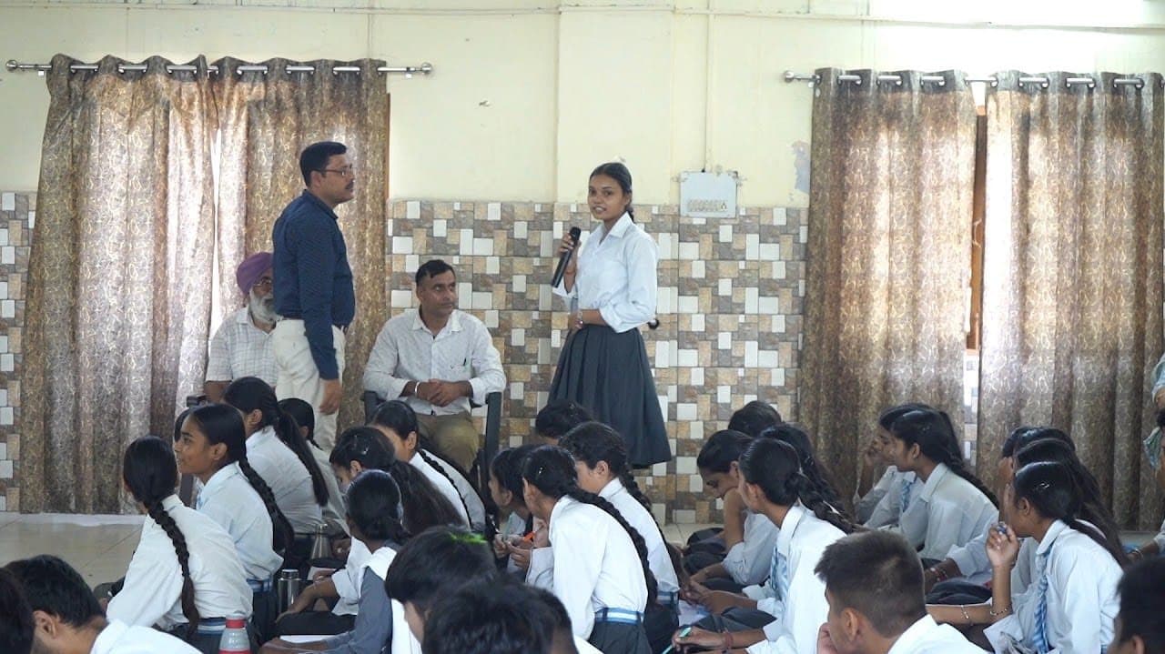 Students seated at desks in a classroom while a teacher stands with a microphone at the side.