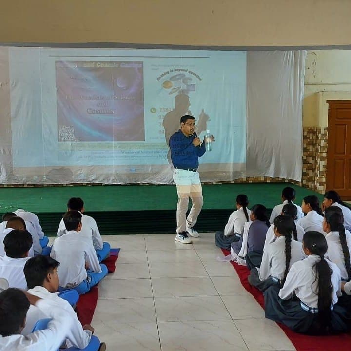 A speaker with a microphone addressing rows of students seated on the floor of a hall.