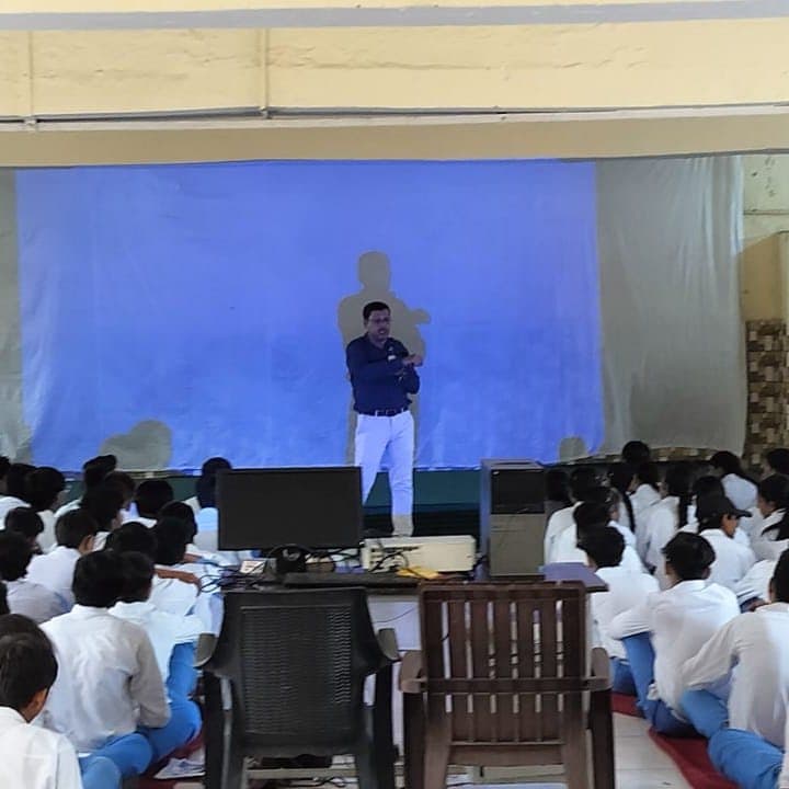 A speaker standing before a large blue projection screen in a darkened hall of seated students.