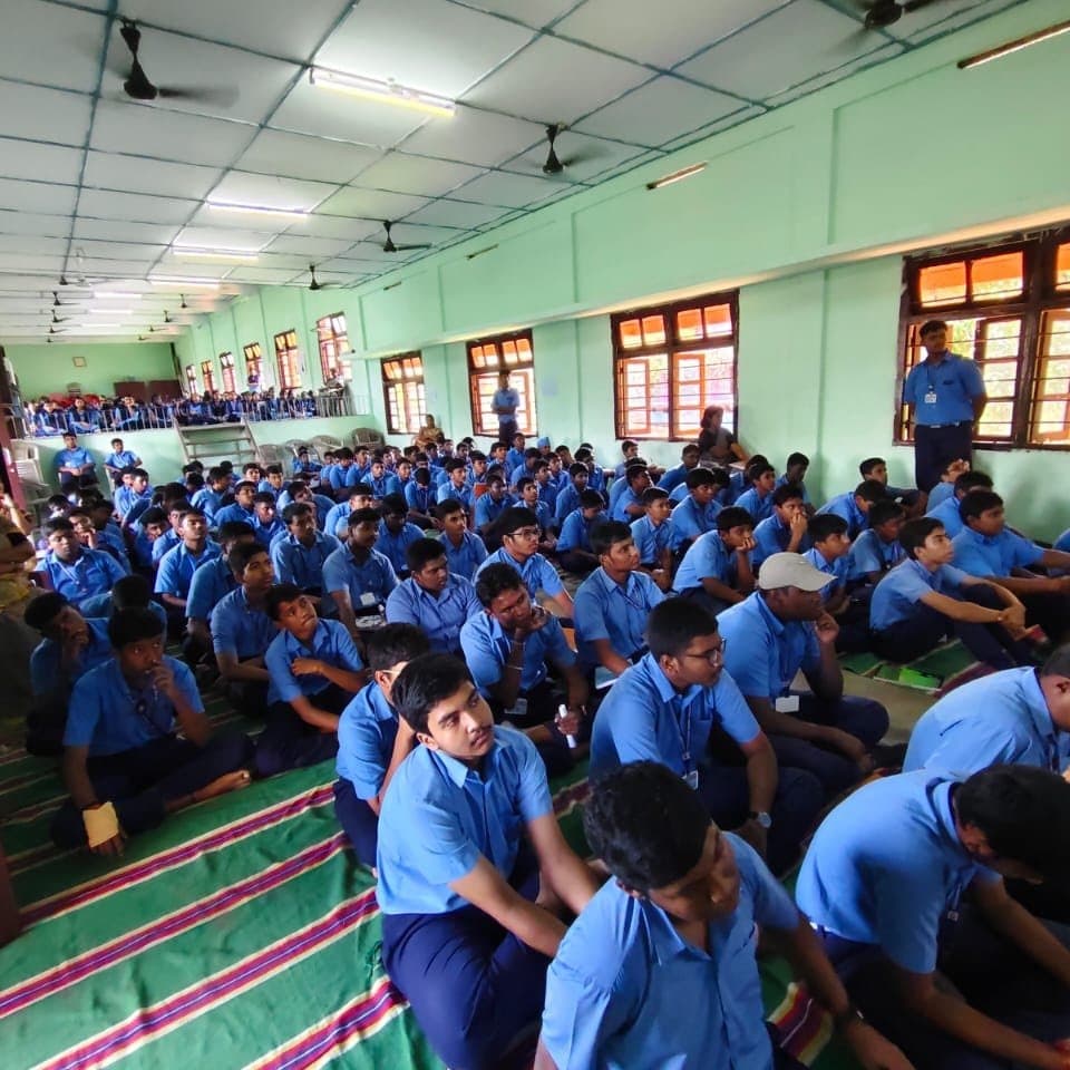 A large group of students in blue uniforms seated on mats in a school hall.
