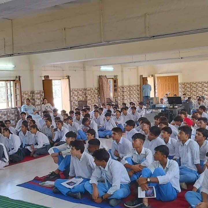 Students seated on mats on the floor of a classroom, some holding printed material, watching a presentation.