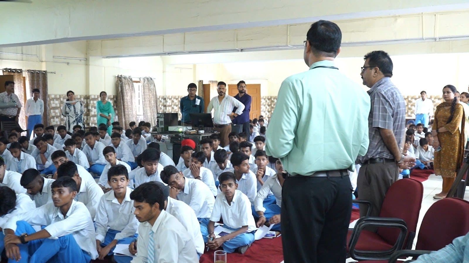 A hall of seated students with facilitators and staff standing along the side.