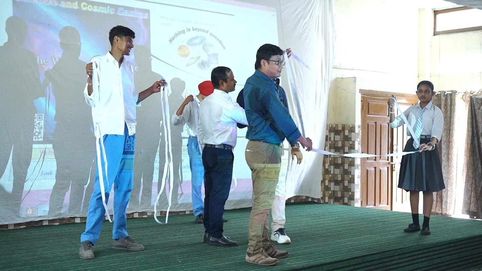 Students on a stage holding long white paper strips while a facilitator directs them.
