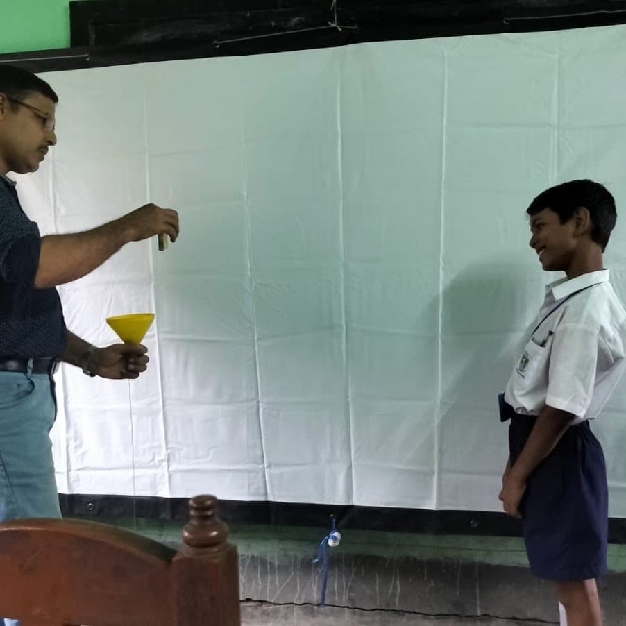 A facilitator holding a yellow funnel at the front of a classroom while a student in uniform watches from across the room.