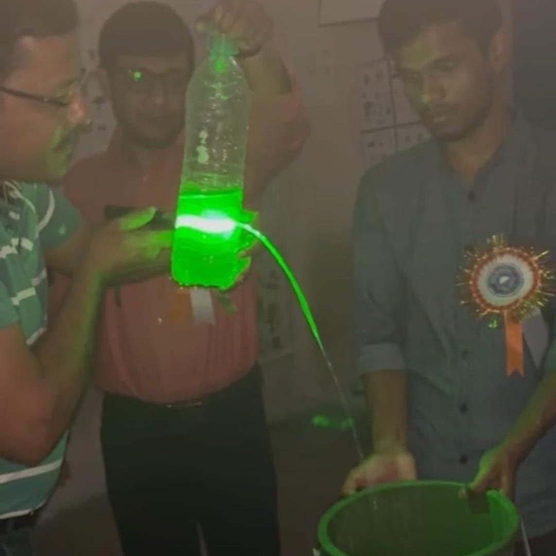 A green laser beam curving along a stream of water pouring from a plastic bottle into a bowl, watched closely by two people.