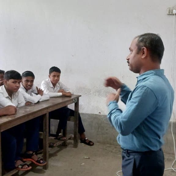 A facilitator gesturing with both hands as he explains something to two schoolboys seated at a desk.