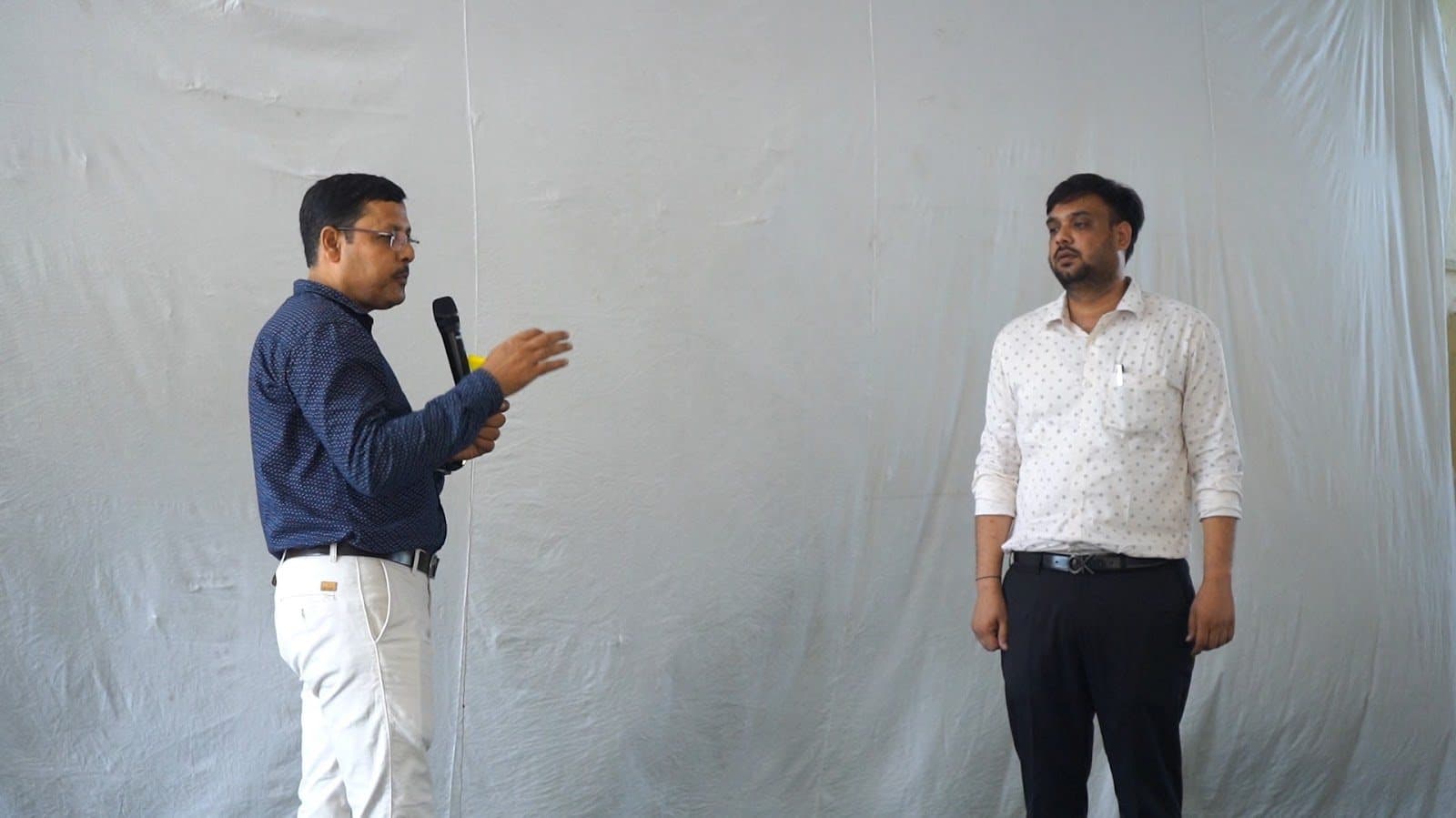 Two facilitators standing at the front of a room against a white backdrop, one holding a microphone.