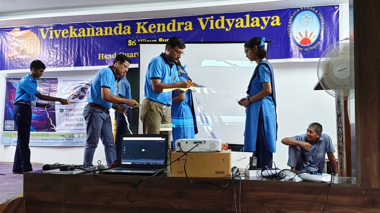 Students in blue uniforms at a table with a laptop and projector, beneath a banner reading Vivekananda Kendra Vidyalaya.