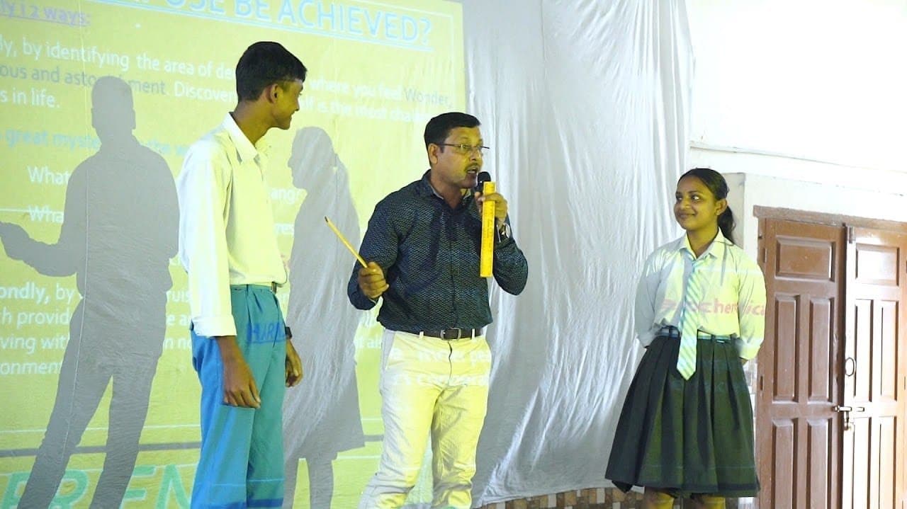 A facilitator holding a microphone beside a student on a stage, a projected slide behind them.