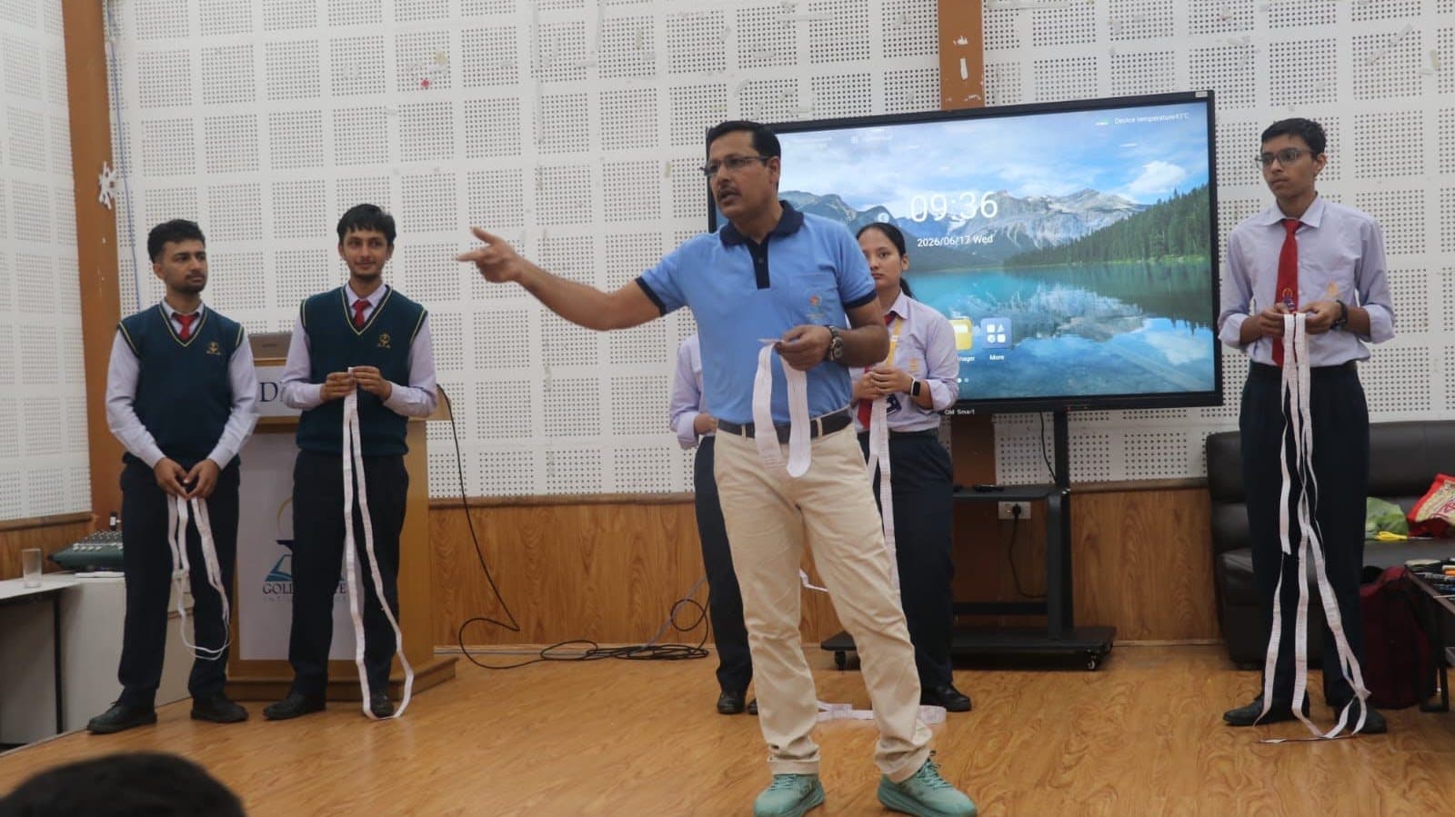 A speaker gesturing on a school stage while two students hold long white strips, a projected landscape behind them.