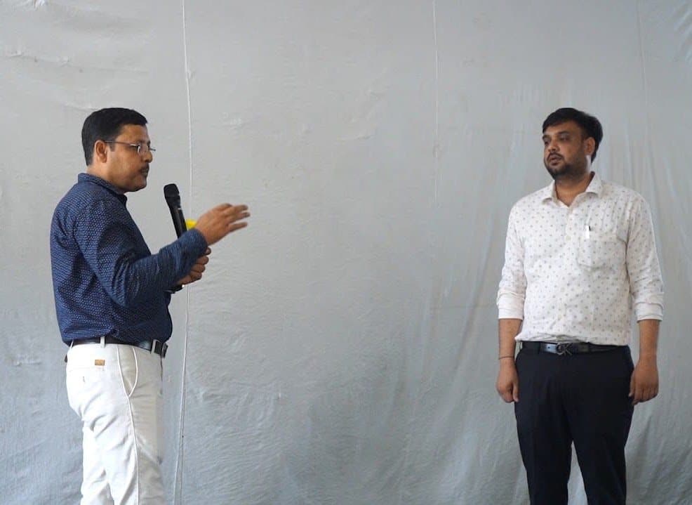 Two facilitators at the front of a room against a white backdrop, one speaking into a microphone.
